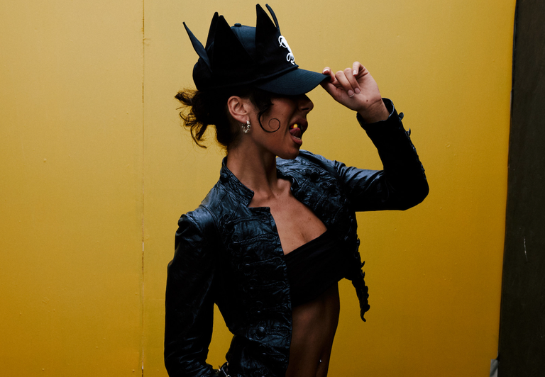 Musician Miss Kaninna posing against a bright yellow backdrop, wearing a black leather jacket and black crown-like hat.